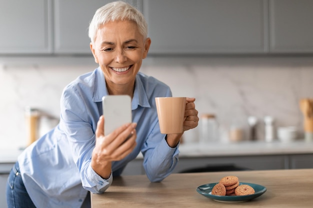 Relaxed mature lady holding smartphone and cup of coffee indoor