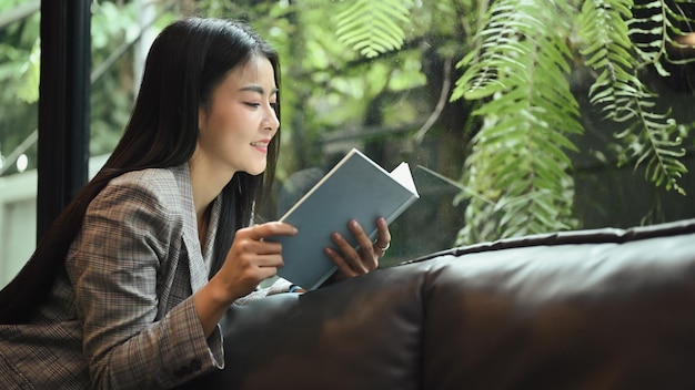 Relaxed millennial businesswoman taking a break from work and reading a book on sofa