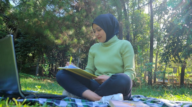 Relaxed muslim woman enjoying weekend at park sitting on grass and writing book empty space