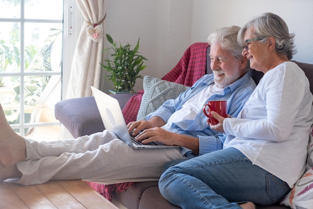 Relaxed senior couple at home sitting on sofa barefeet using laptop. Two elderly retirees enjoying free time and technological devices, social media