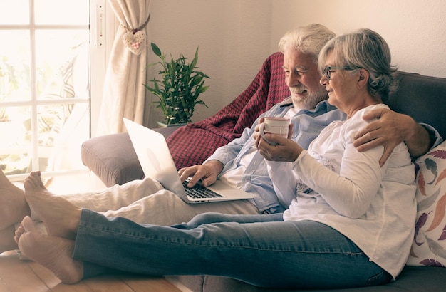 Relaxed senior couple lying on sofa at home using laptop Two elderly retirees enjoying free time and technological devices woman holding in hand a cup of coffee or tea