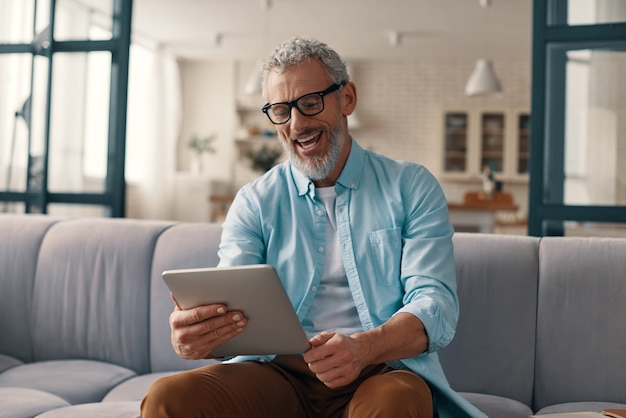 Relaxed senior man using digital tablet and smiling while sitting on the sofa at home