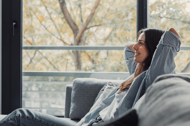 Photo relaxed serene pretty young woman feel fatigue lounge on comfortable sofa hands behind head rest at home happy calm lady dream enjoy wellbeing breathing fresh air in cozy home modern living roomxa