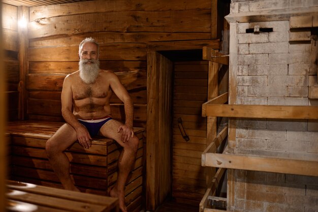 Relaxed thoughtful grey-haired senior man rest at bath house with oak broom. Wellness spa procedure