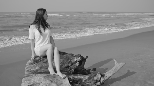 Relaxed woman sitting on log and looking away Female tourist resting on sandy beach near waving sea