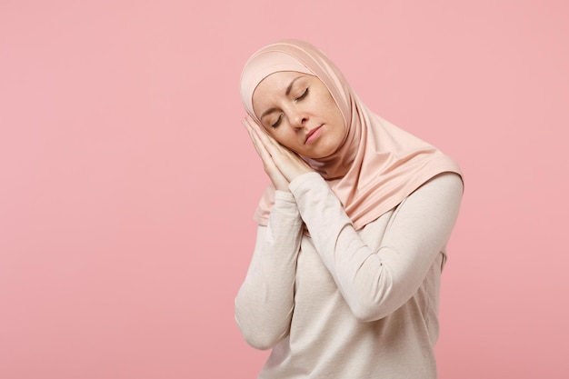 Relaxed young arabian muslim woman in hijab light clothes posing isolated on pink background in studio. People religious lifestyle concept. Mock up copy space. Sleeping with folded hands under cheek.