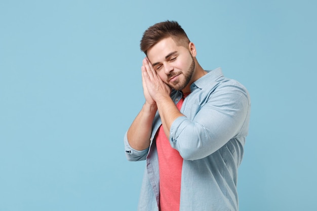 Relaxed young bearded guy in casual shirt posing isolated on pastel blue background studio portrait. People sincere emotions lifestyle concept. Mock up copy space. Sleep with folded hands under cheek.