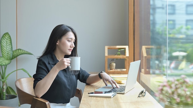 Relaxed young business woman drinking coffee and checking email on her laptop computer