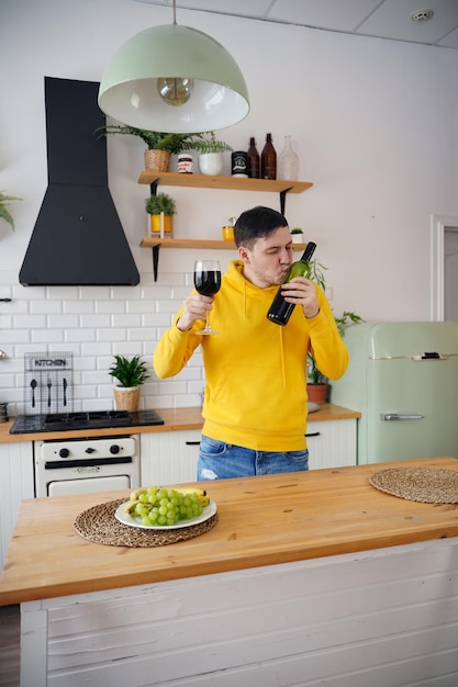 Relaxed young man kisses bottle of red wine standing at kitchen table Adult guy resting with alcohol in kitchen