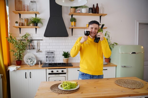 Relaxed young man poses with bottle and glass of red wine in kitchen Adult happy guy presses alcohol to face