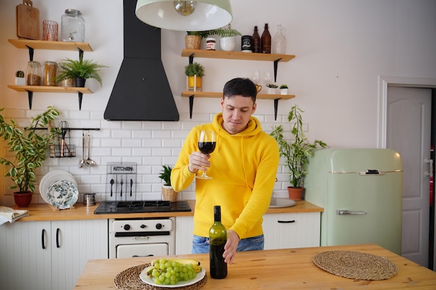 Relaxed young man poses with glass of red wine standing at kitchen table Adult guy resting with alcohol and fruits in kitchen