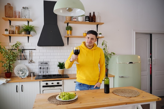 Relaxed young man poses with glass of red wine standing at kitchen table Adult guy resting with alcohol and fruits in kitchen