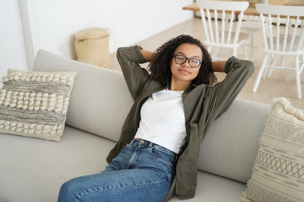 Relaxed young mixed race girl sitting on cozy comfortable sofa resting at home enjoying lazy weekend