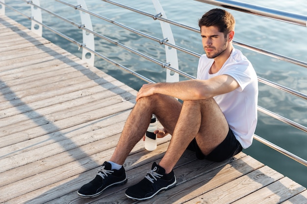 Relaxed young sportsman with bottle of water sitting and relaxing on pier