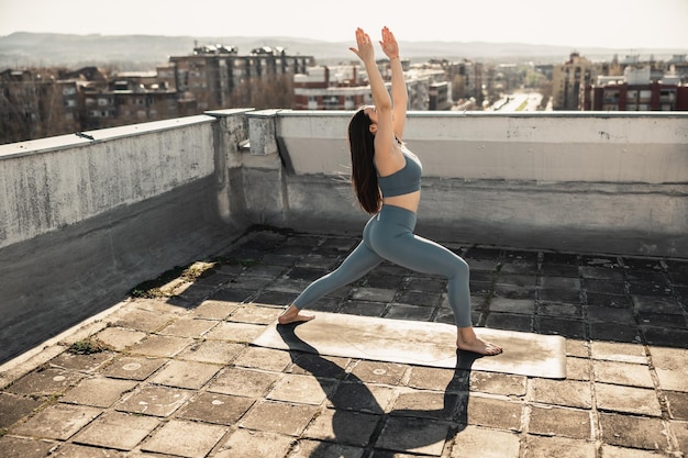 Relaxed young woman doing yoga stretching exercise on a rooftop terrace. City buildings in the background.