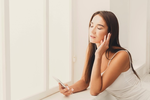 Relaxed young woman listening to music online on smartphone. Girl in earphones enjoying favorite playlist while standing near the window at home