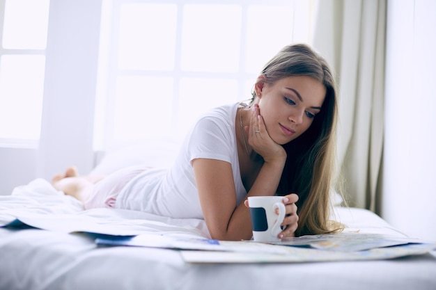 Relaxed young woman sitting on bed with a cup of coffee and digital tablet