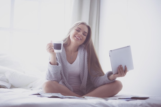 Relaxed young woman sitting on bed with a cup of coffee and digital tablet