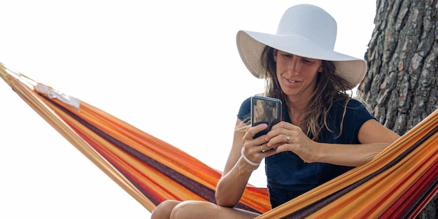 Relaxed young woman with sunhat sitting in hammock browsing on her mobile phone.