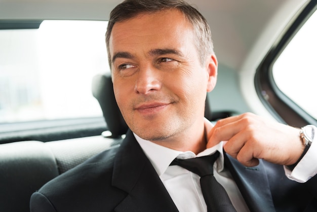 Relaxing after business trip. Satisfied mature businessman adjusting his necktie while sitting on the back seat of a car