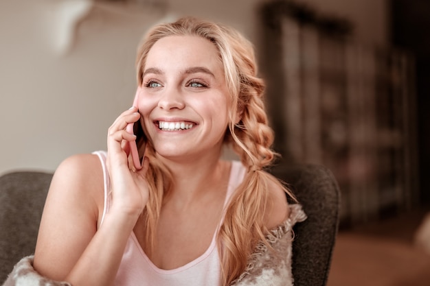 Relaxing in apartments. Cheerful blonde woman with lush braids laughing during conversation and showing her white teeth