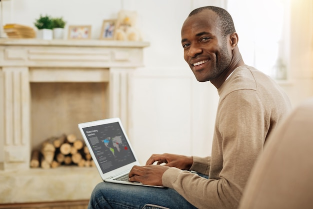 Relaxing. Gleeful dark-eyed afro-american man smiling and working on the laptop while sitting on the couch and a fireplace in the background