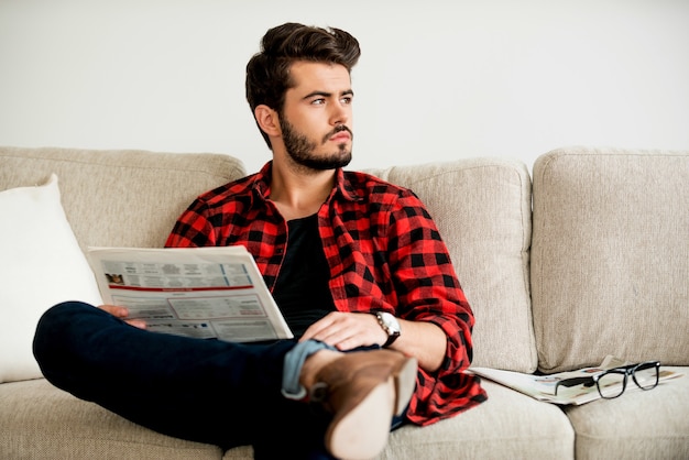 Relaxing on sofa. Thoughtful young man holding newspaper and looking away while sitting on sofa