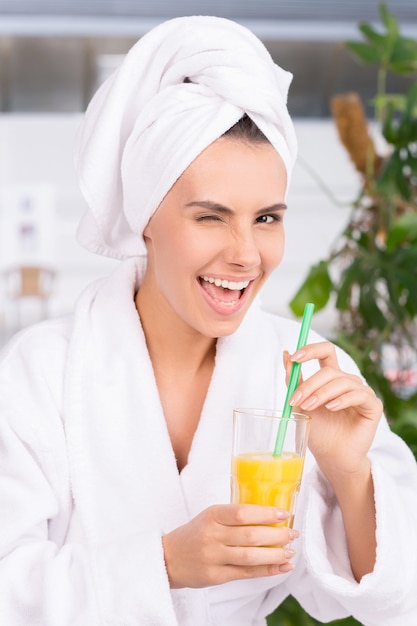 Relaxing at spa. Beautiful young woman in bathrobe holding s glass with juice and winking to you