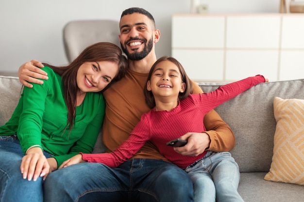 Relaxing Together. Portrait of happy young Arab family of three people watching television, parents sitting with their daughter on comfortable sofa in modern living room, laughing at the comedy