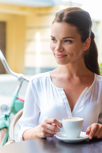 Relaxing with cup of fresh coffee. Beautiful young woman drinking coffee and looking away while sitting at the sidewalk cafe