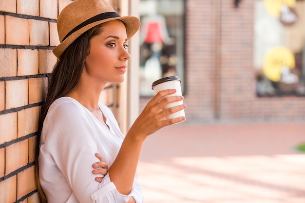 Relaxing with cup of fresh coffee. Side view of thoughtful young woman in funky hat holding cup with hot drink and looking away while leaning at the wall outdoors