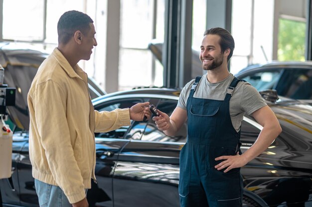 Reliability. Young adult confident mechanic taking car key from black smiling man standing in workshop during day