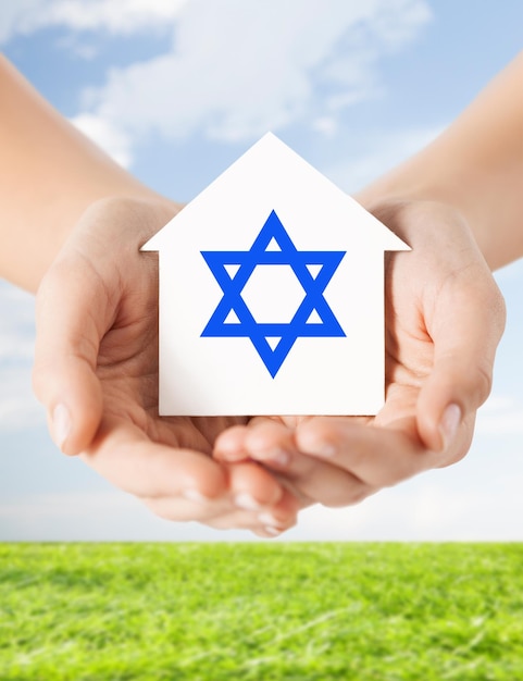 religion, judaism and charity concept - close up of woman hands holding house with star of david over blue sky with grass background