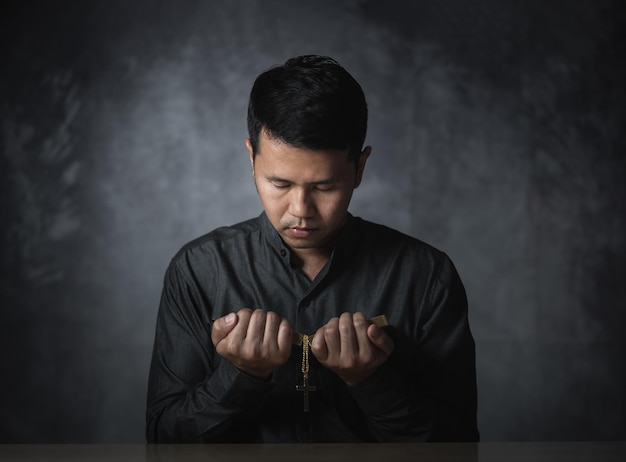 Religious asian man praying holding necklace of crosses and bible verses praying for holy blessings sitting on the table gray background Spirituality and religion Christian prayer religion concept