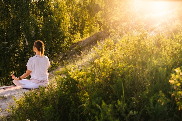 Remote back view of young woman meditating in lotus position sitting on top of rock