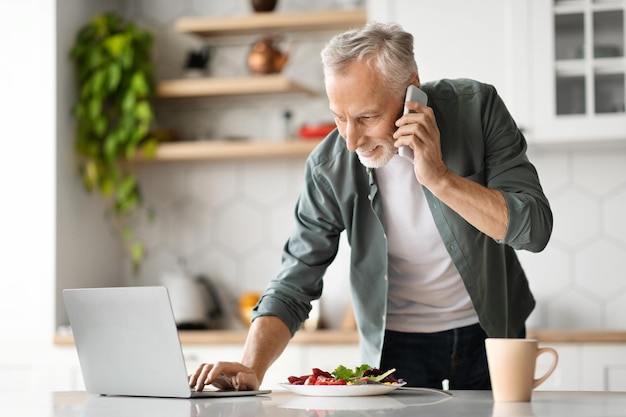 Remote business senior man using cellphone and working on laptop in kitchen