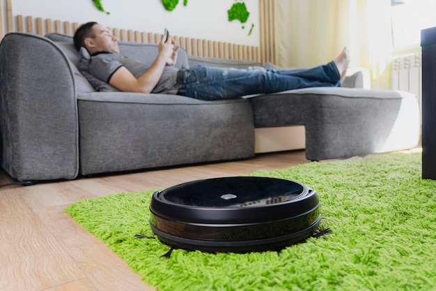 Remote control of robot vacuum cleaner via smartphone Smart technologies at home Internet of Things concept Robot vacuum cleaner cleaning the living room man enjoy rest on sofa at home