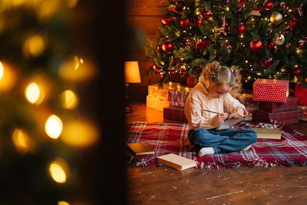 Remote view of focused charming little blonde curly child girl writing letter to santa claus sitting