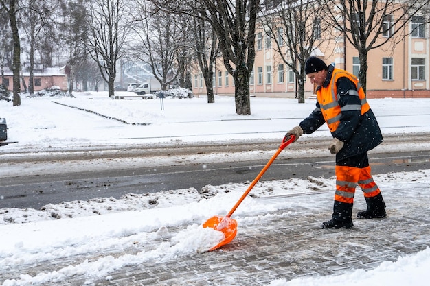 Removing snow from the sidewalk after snowstorm A road worker with a shovel in his hands and in special clothes cleans the sidewalk and the road from snow Snowstorm and hurricane in the city