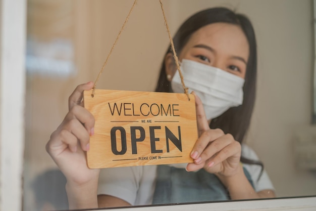 Reopen asian young owner retail coffee shop woman turning sign board open wearing face mask protect to pandemic of coronavirus reopen store after close lockdown quarantine in covid19 new normal