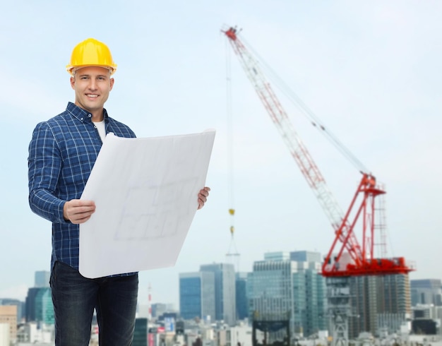 repair, construction, building, people and maintenance concept - smiling male builder or manual worker in helmet with blueprint over city construction site background