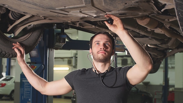 Repairman of Car Workshop Is Standing Under Lifted Automobile and Examining Details.