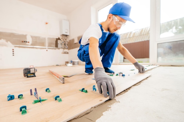 Repairman in grey gloves and uniform laying tiles with tile leveling system on the floor in house