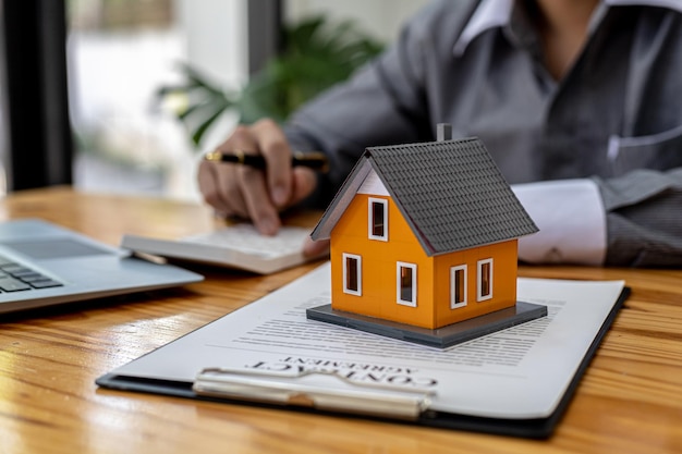 Replica of a small orange house sits on a table, a housing project salesperson is drafting a sales contract for a customer who reserves a house in the project he maintains. Real estate trading concept