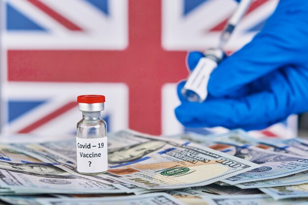 Researcher hand in blue gloves holding covid-19 vaccine against background of flag of England and money disease preparing for human clinical trials vaccination shot, medicine concept.