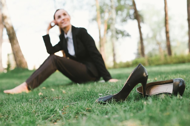 Rest after work in the office Beautiful young business woman in a black suit is sitting in the park on the lawn and smiling Women's shoes in the foreground