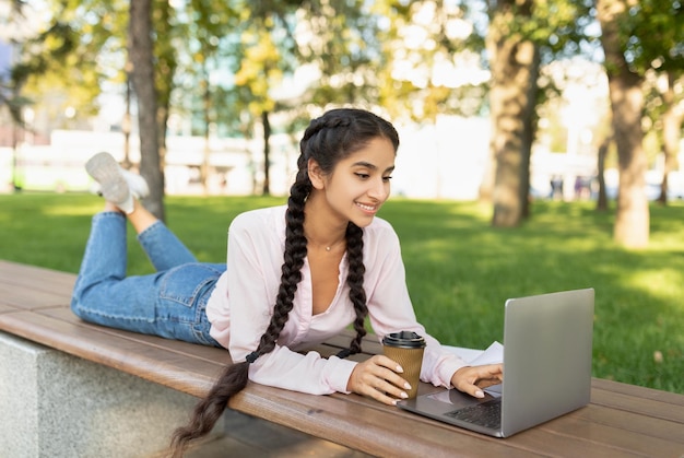 Rest at campus happy indian college student girl relaxing outdoors with coffee and laptop lying on