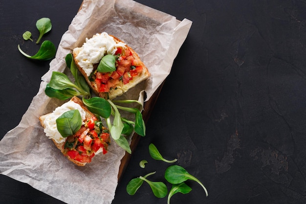 Restaurant appetizer on black background. Crusty bruschetta with concasse tomatoes, stracciatella cheese decorated with spinach. Delicious and healthy meals, top view, copy space