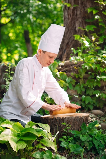 A restaurant chef in uniform rolls out dough on a stump in the woods.