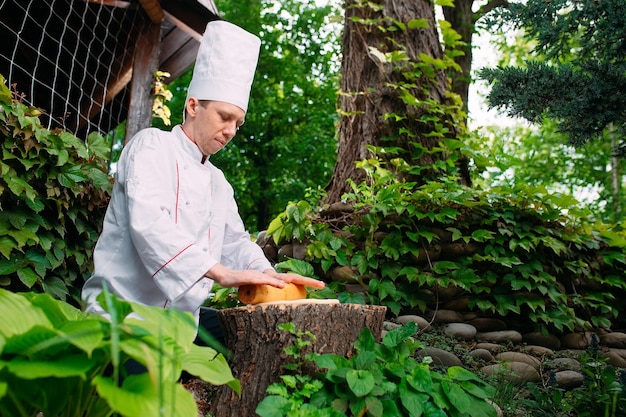 A restaurant chef in uniform rolls out dough on a stump in the woods.